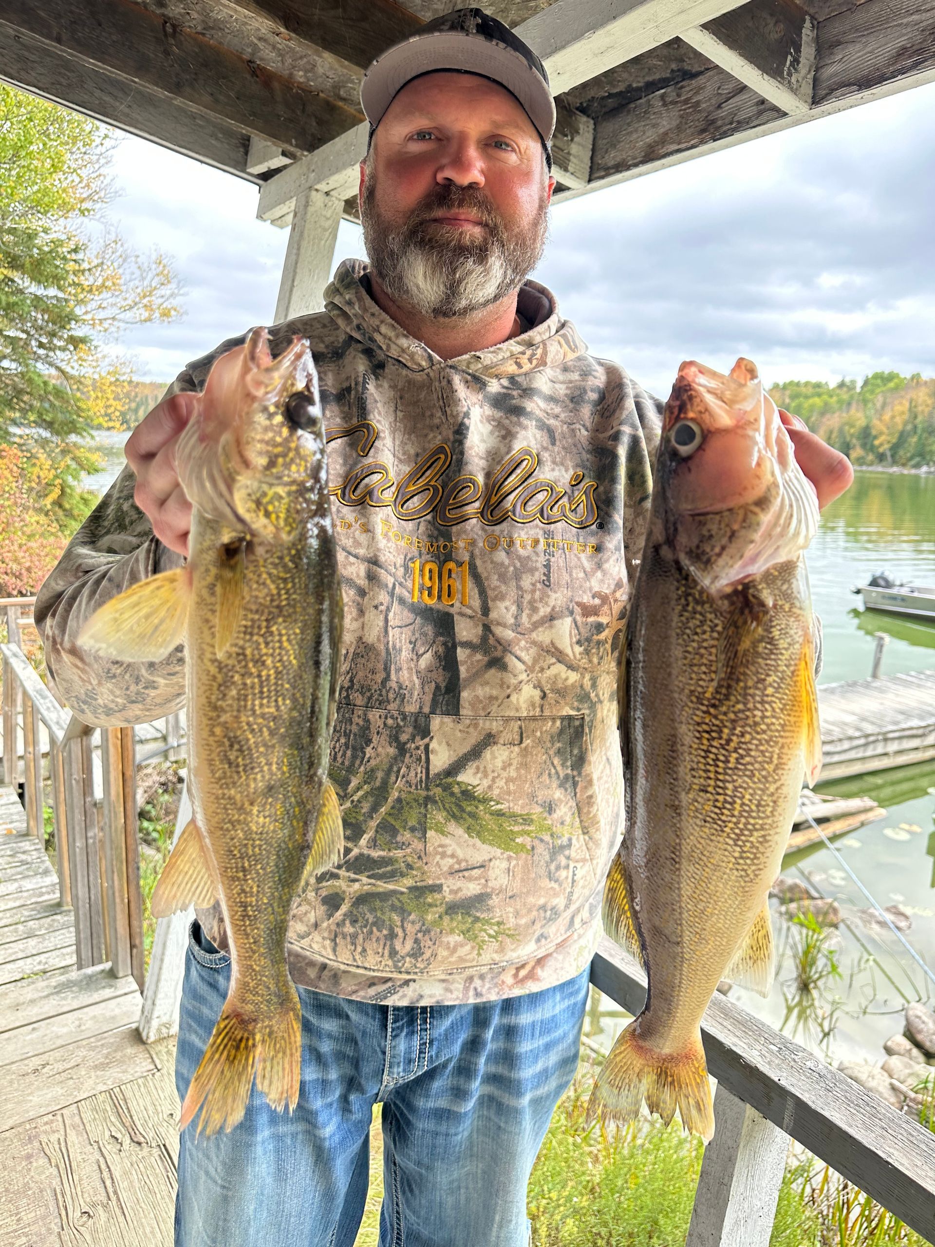 Man holding two large fish on a dock, wearing camouflage hoodie and cap, with water and trees behind him