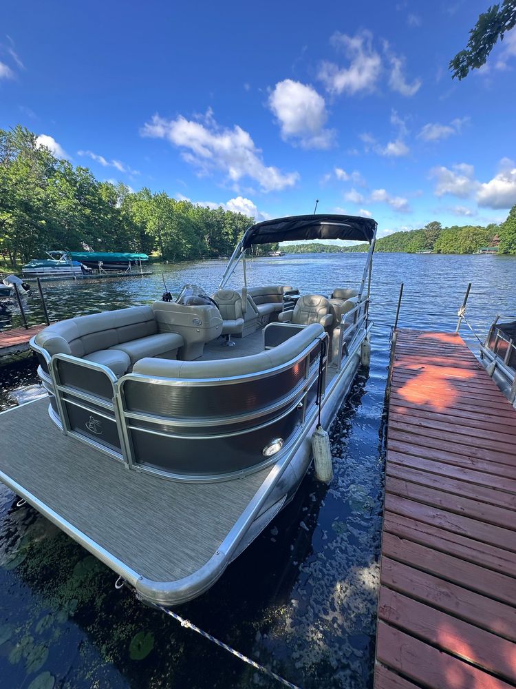 Pontoon boat docked on a calm lake, with green forested shoreline and overcast sky.