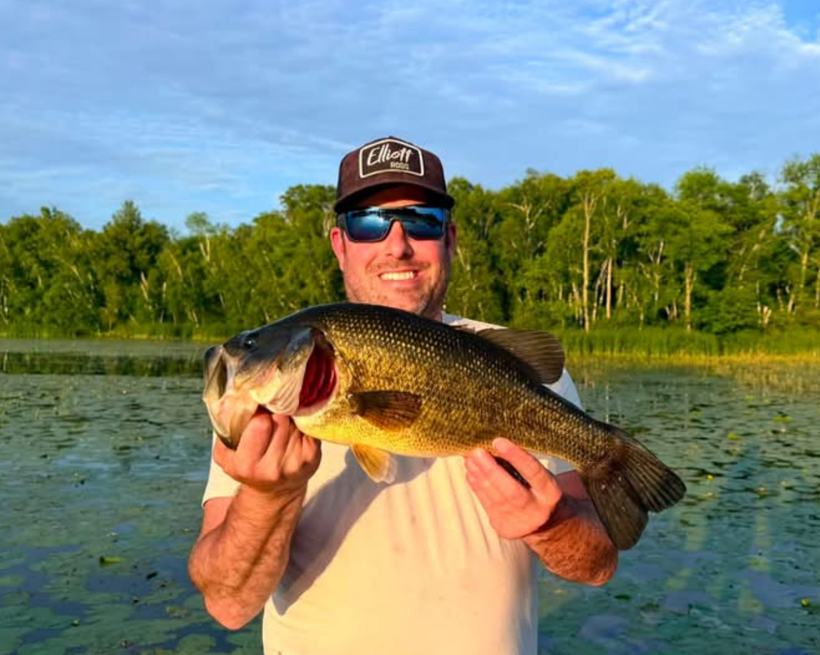 Person holding a large fish on a boat on a lake, wearing a tan jacket, jeans, and a red cap.