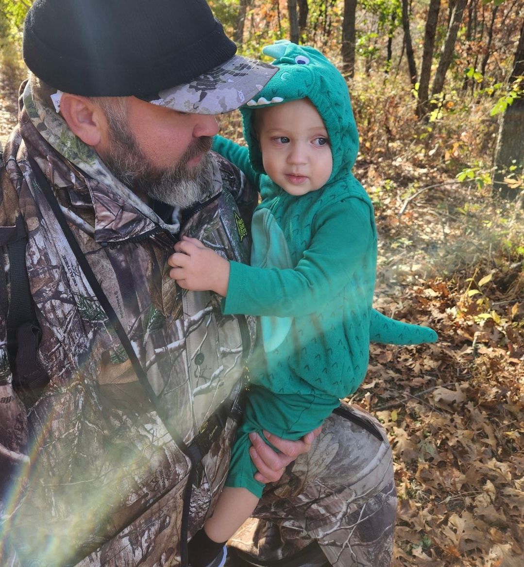 Adult in camouflage holding a toddler in a teal dinosaur costume in a wooded autumn setting
