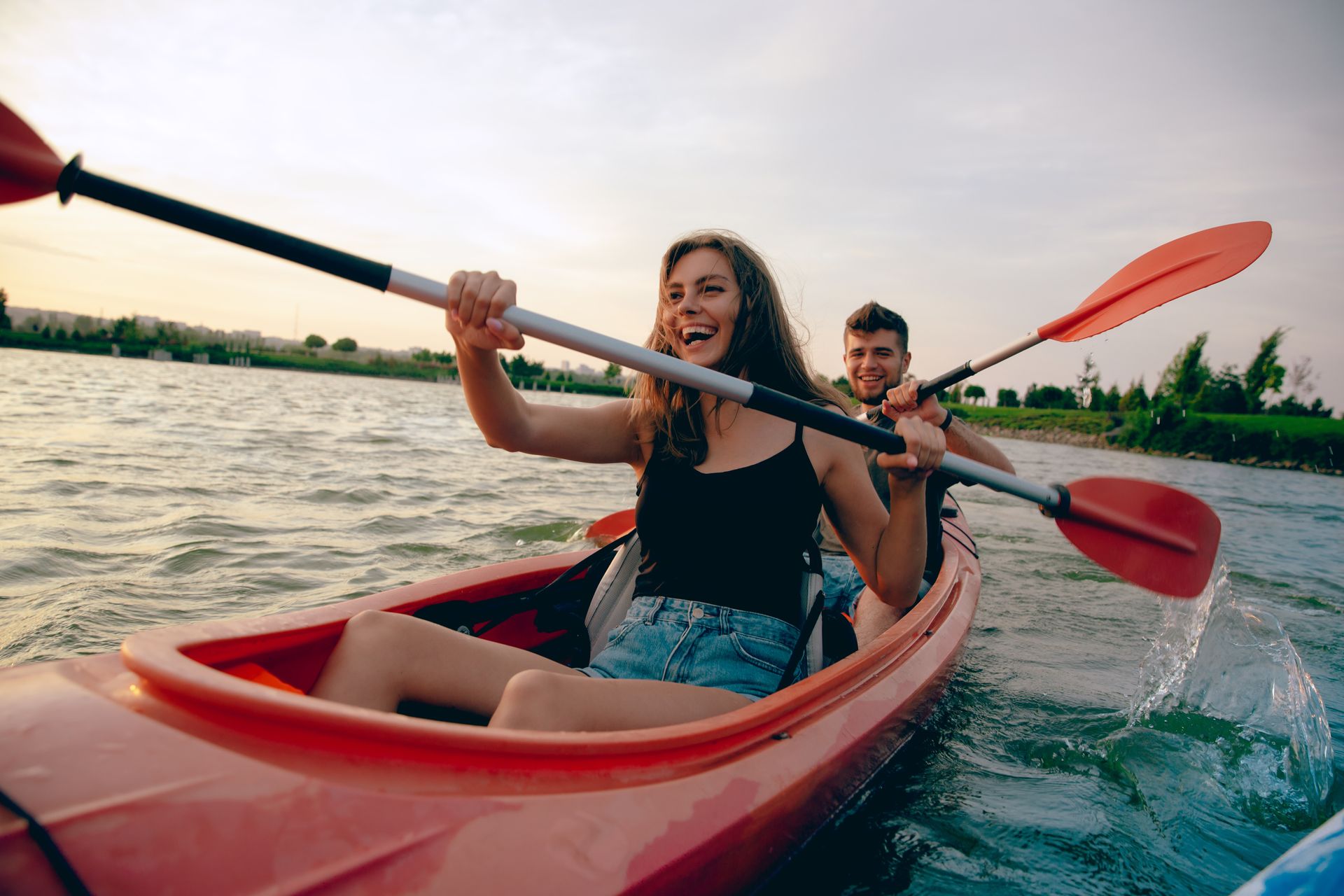 Two people kayaking on a river at sunset, splashing water with red paddles in a red kayak.
