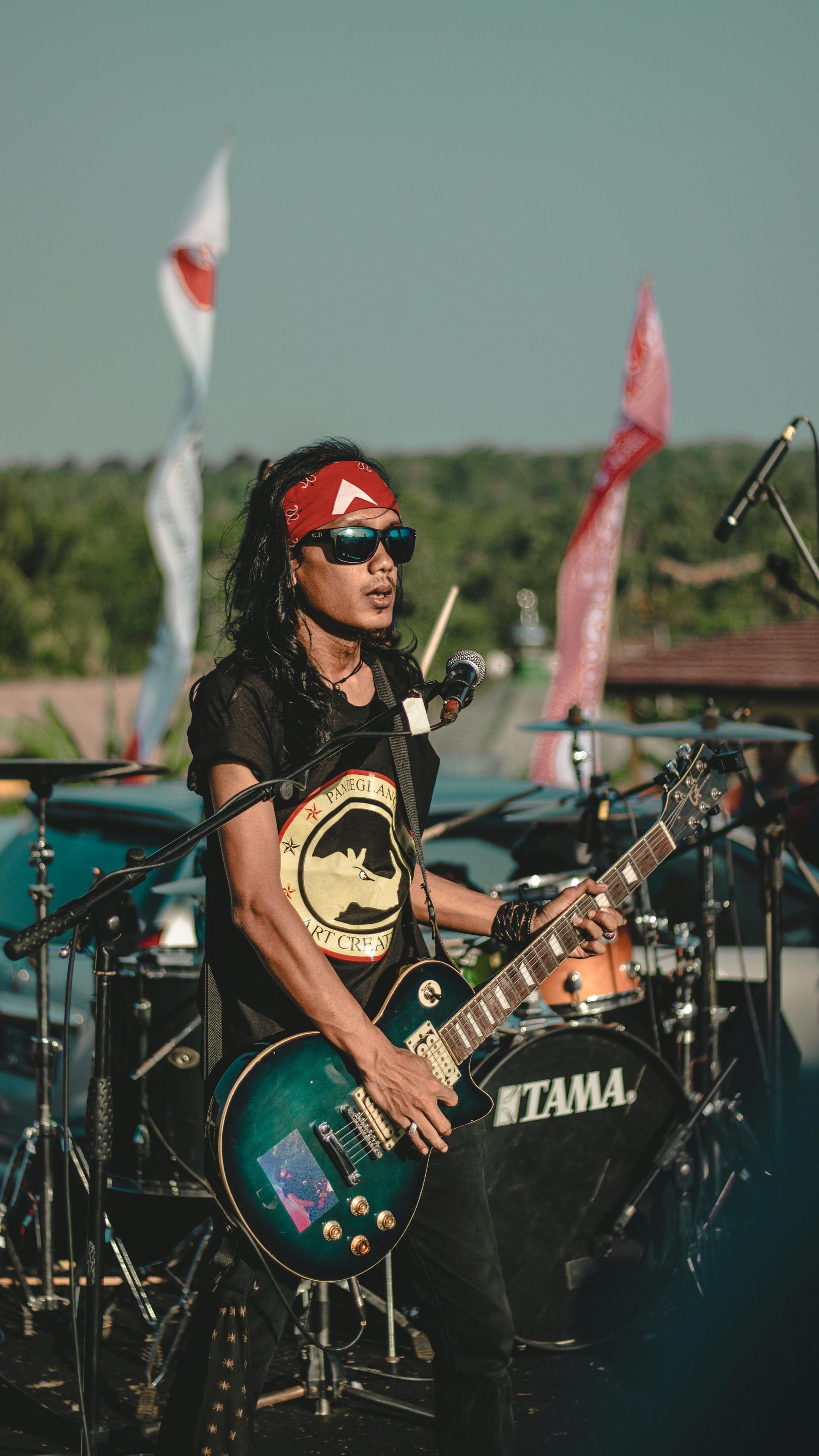 Musician playing a black electric guitar onstage, wearing sunglasses and a red bandana, with drums behind.