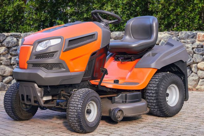 Orange and gray lawn tractor with seat, parked on a gravel surface in front of a hedge