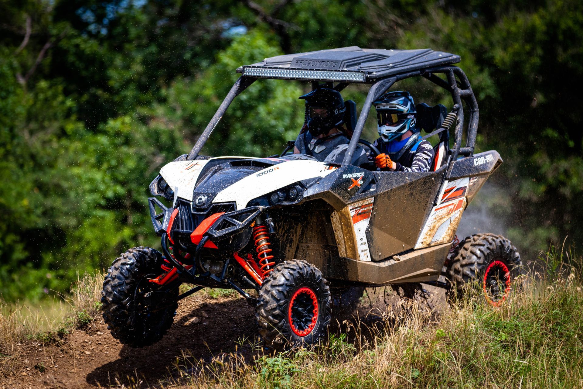 Off-road side-by-side vehicle driving through tall grass on a dirt trail, with a driver wearing a helmet.