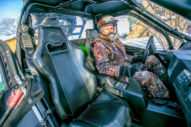 Person in camo gear seated inside a damaged vehicle, viewed from the passenger side.
