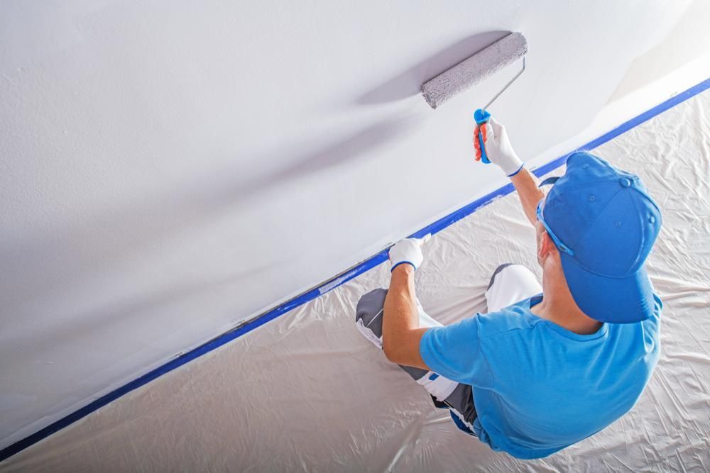 A Man Is Sitting On The Floor Painting A Wall With A Roller — Strait Edge Painting In Tweed Heads, NSW