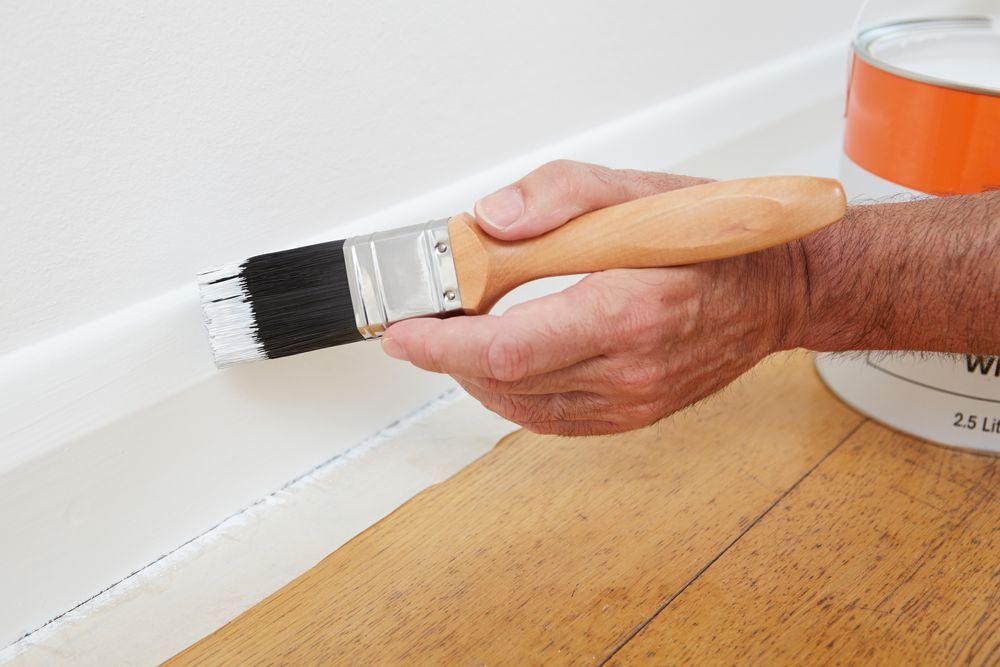 A Hand Uses a Paintbrush to Apply White Paint to a Wooden Baseboard Trim — Strait Edge Painting In Tweed Heads South, NSW