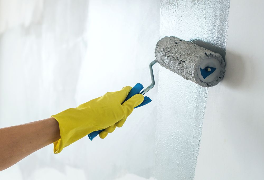 A Gloved Hand Uses a Paint Roller to Apply Grey Paint Onto a White Wall — Strait Edge Painting In Gold Coast, NSW