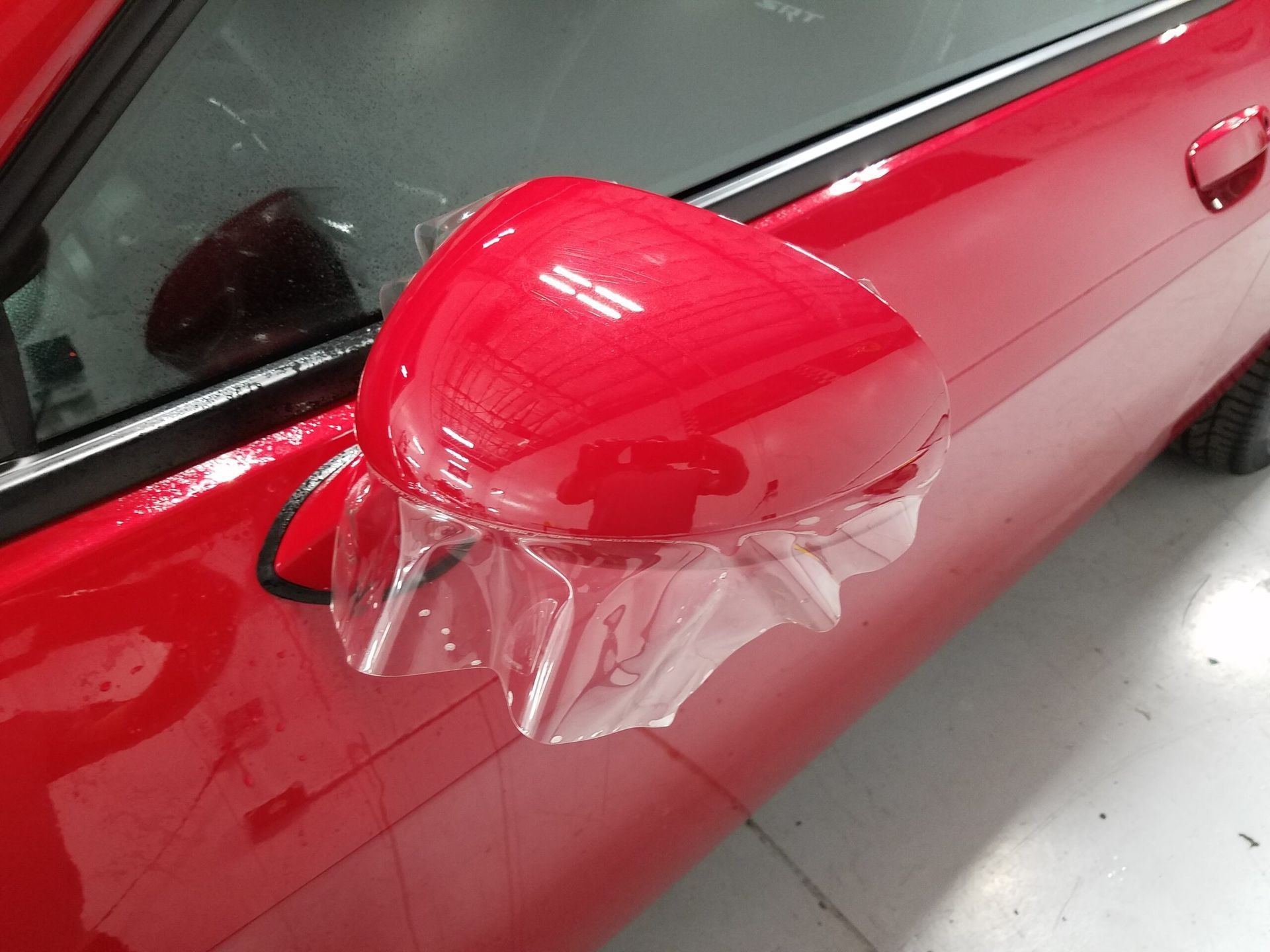 Red car side mirror covered in clear protective plastic wrap.