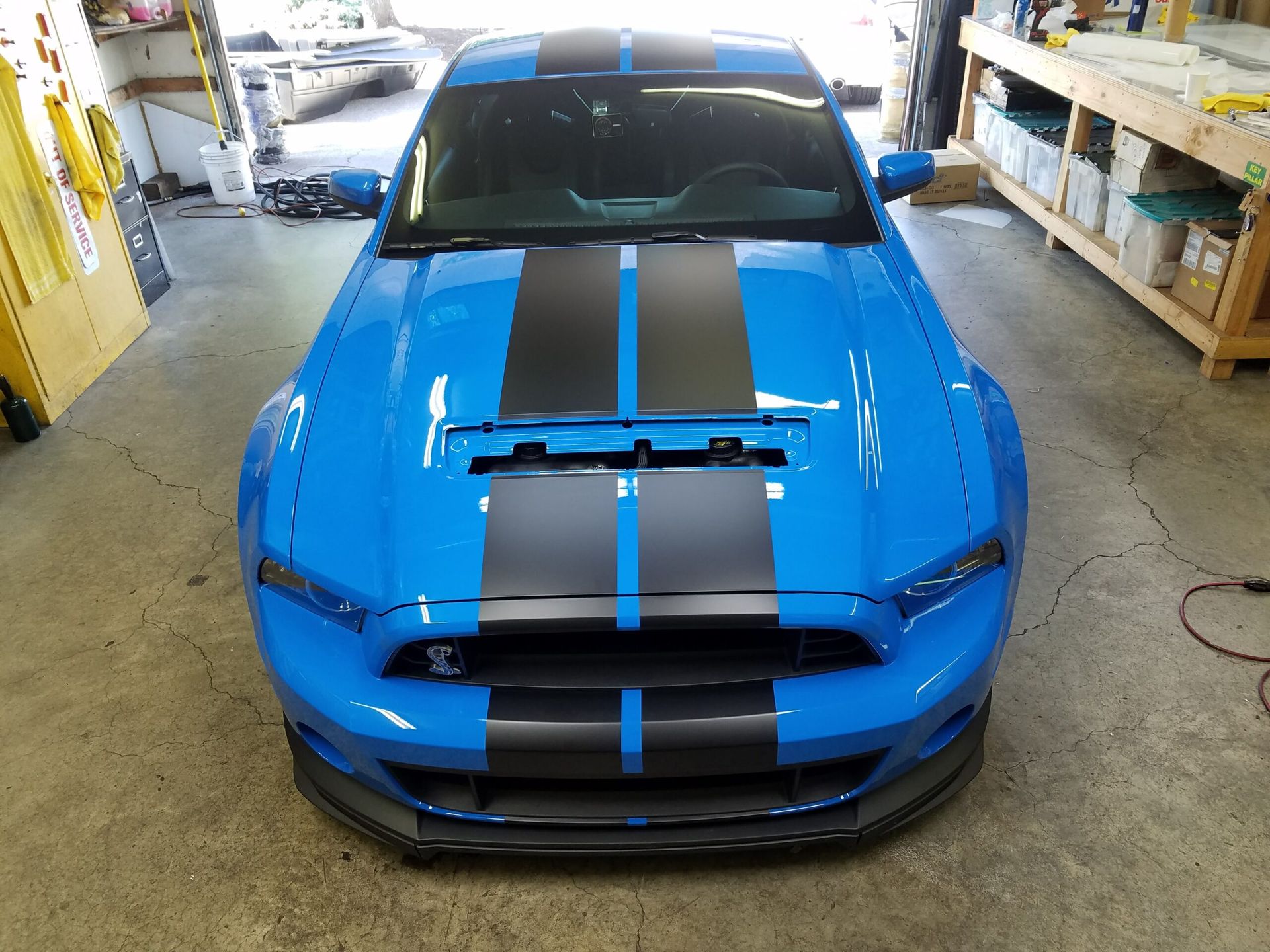 Blue sports car with black racing stripes parked indoors in a garage or workshop.