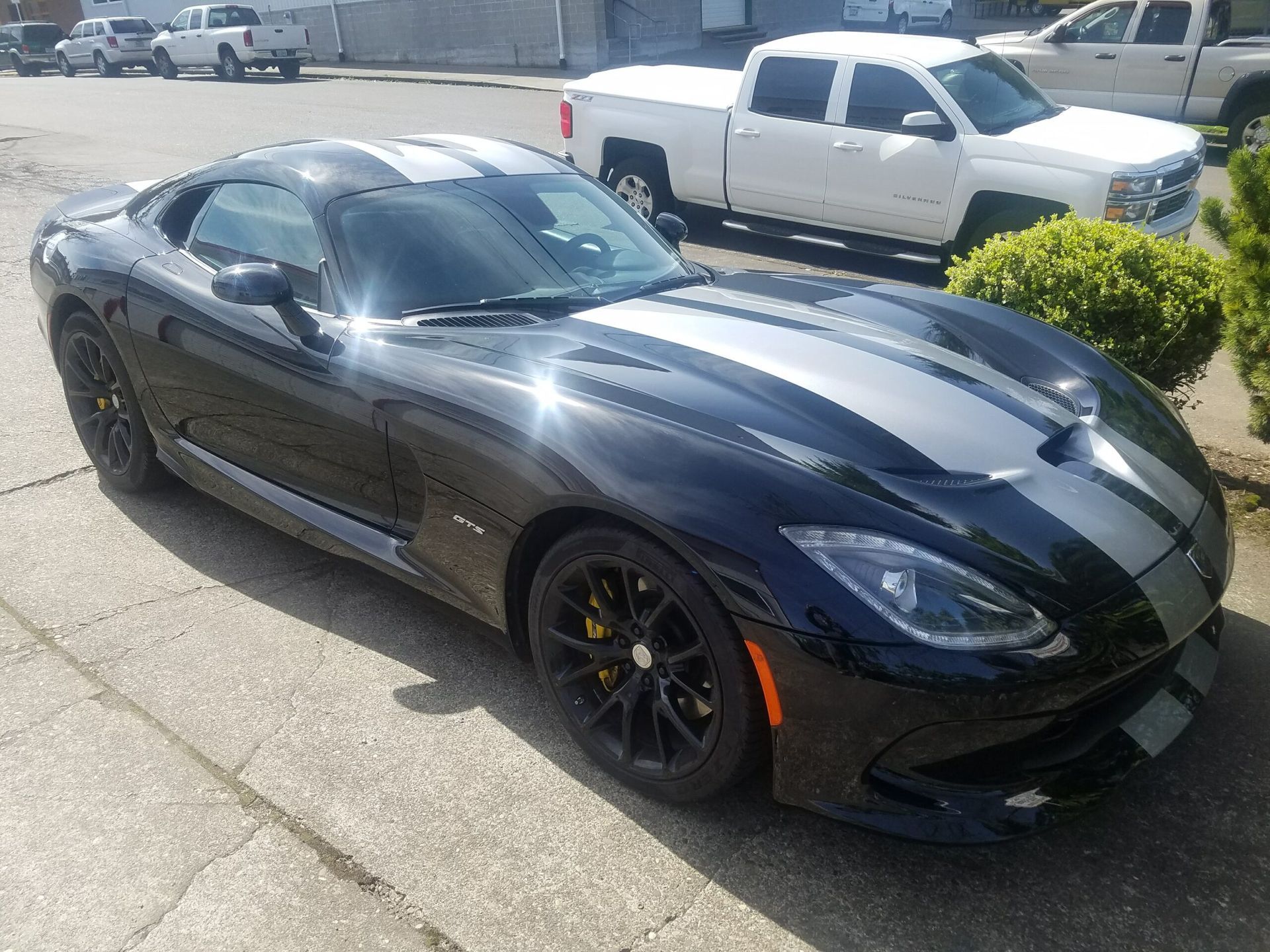 Black sports car with silver racing stripes parked on a sunny street near a white pickup truck.