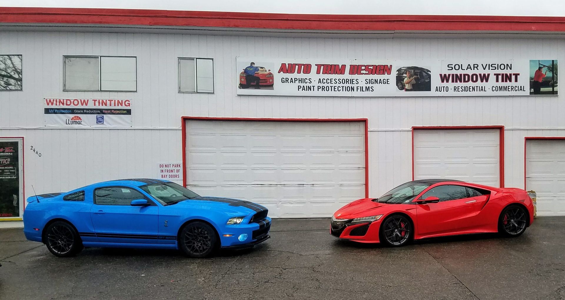Two sports cars, blue and red, parked in front of a window tint shop with garage doors