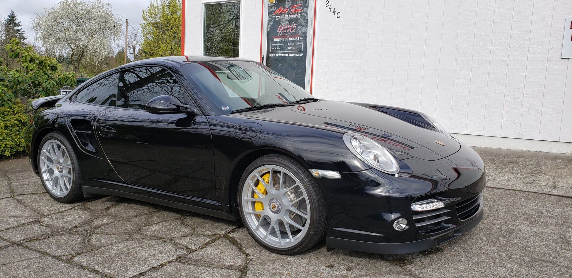 Black sports coupe parked beside a white building, with silver wheels and yellow brake calipers