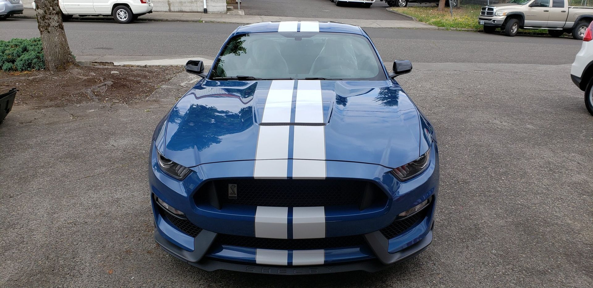 Blue sports car with white racing stripes, front view, parked on gravel in a lot