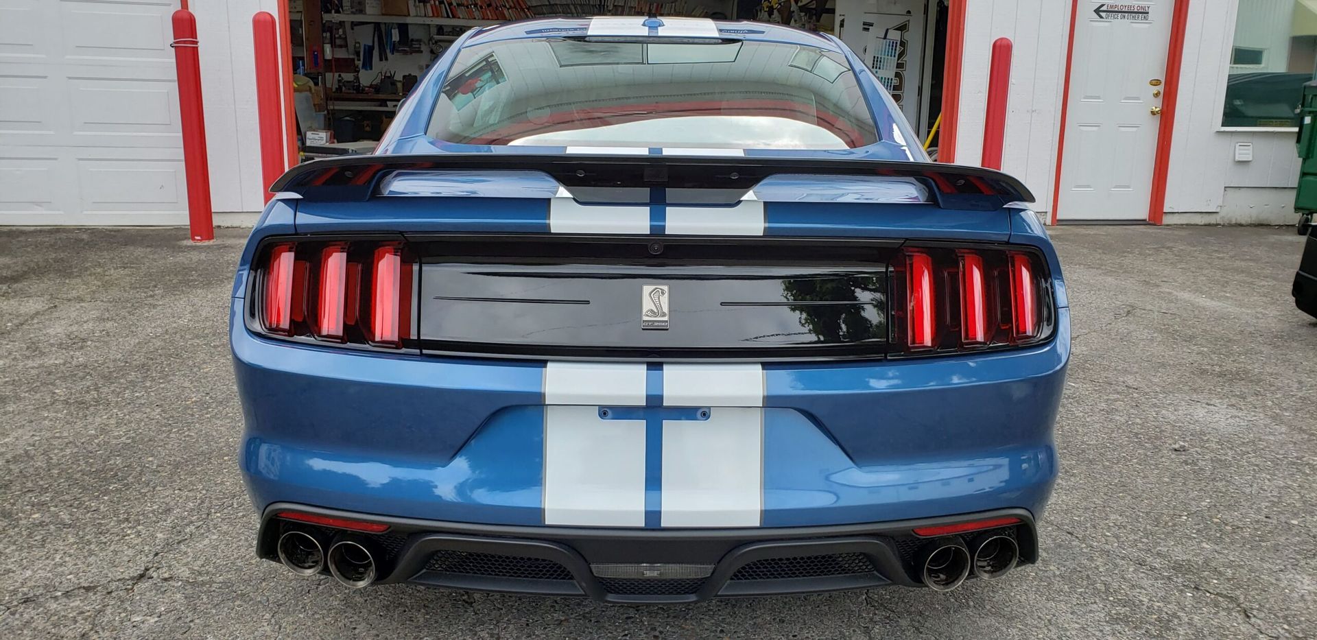 Rear view of a blue Ford Mustang with white racing stripes parked in front of a garage