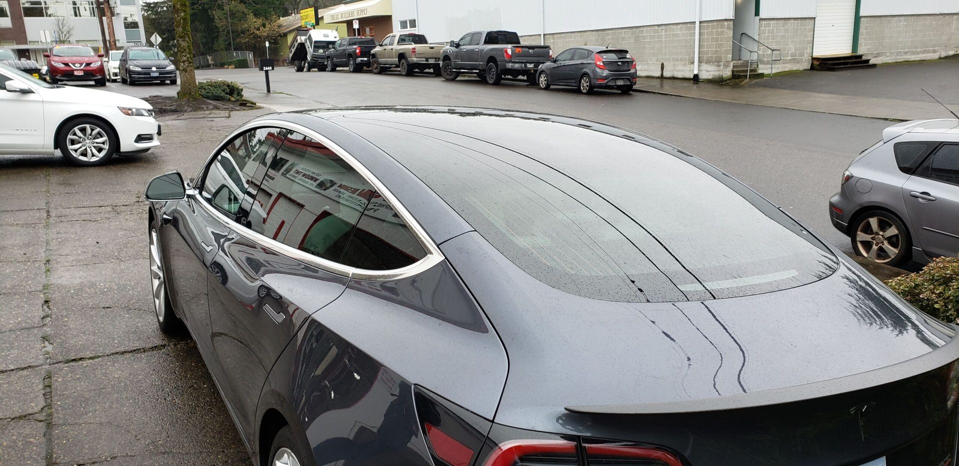 Gray Tesla parked on a wet street, viewed from the rear side near other cars.