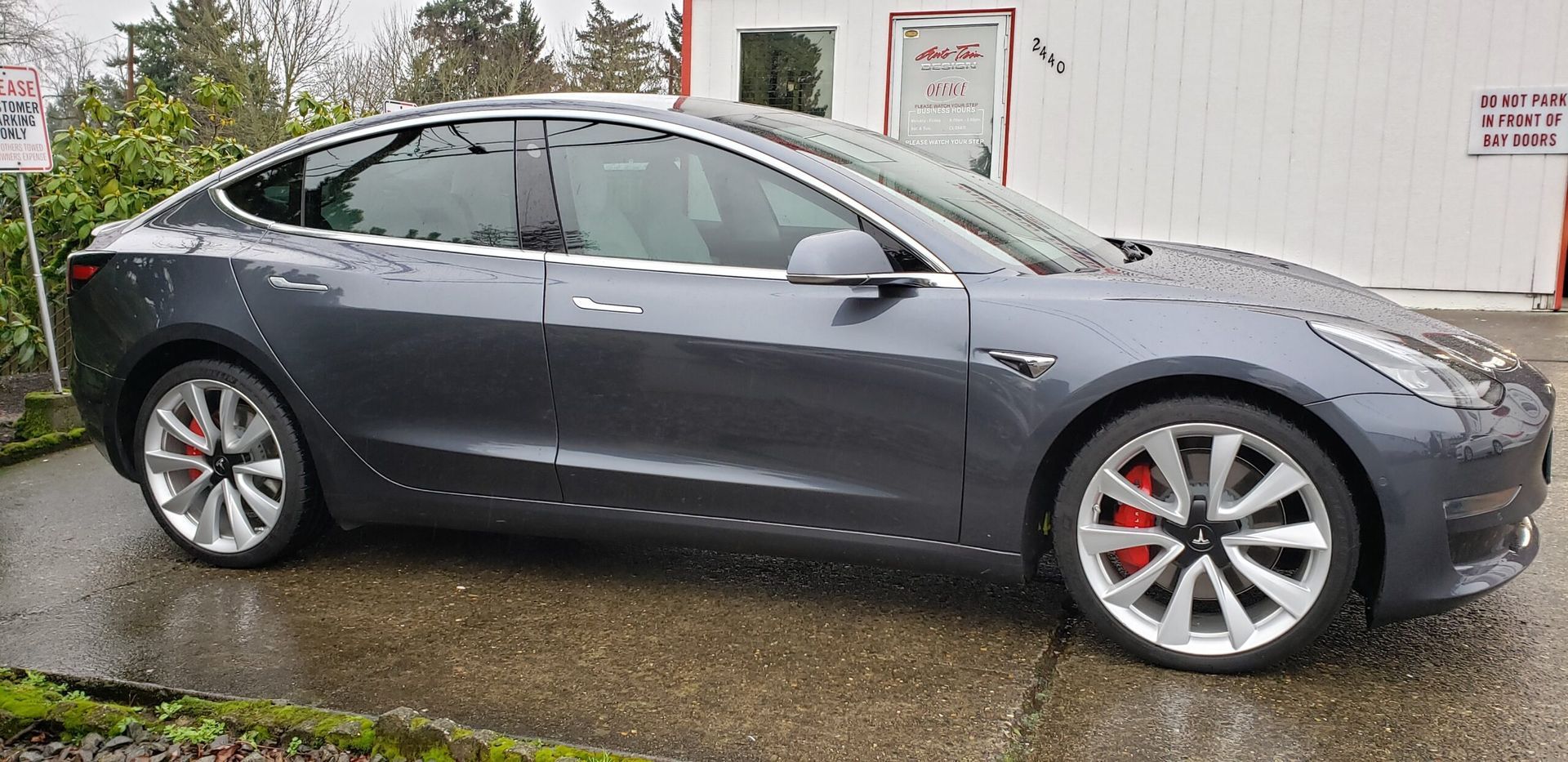 Gray Tesla sedan parked on a wet driveway beside a white building
