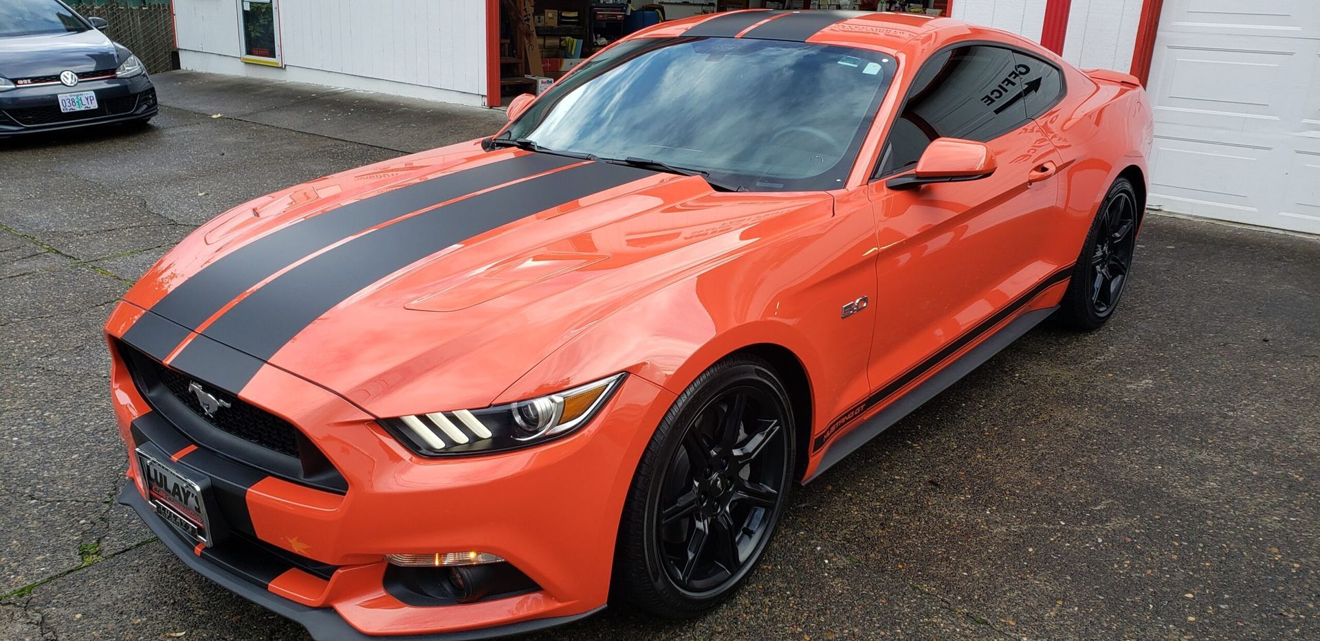 Orange Ford Mustang with black racing stripes parked outdoors on wet pavement