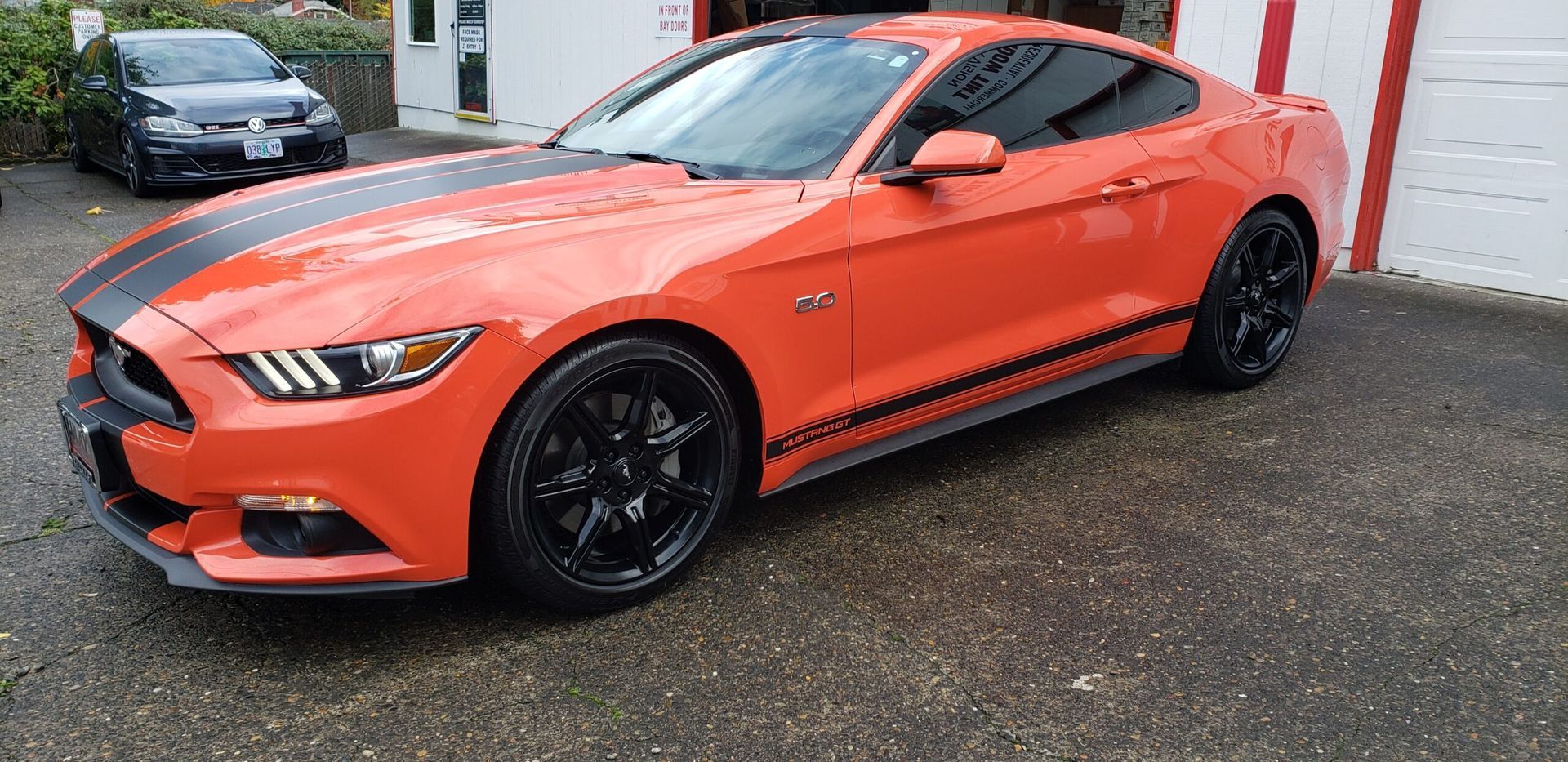Orange sports car parked on wet pavement near a white building, with a black car behind it.