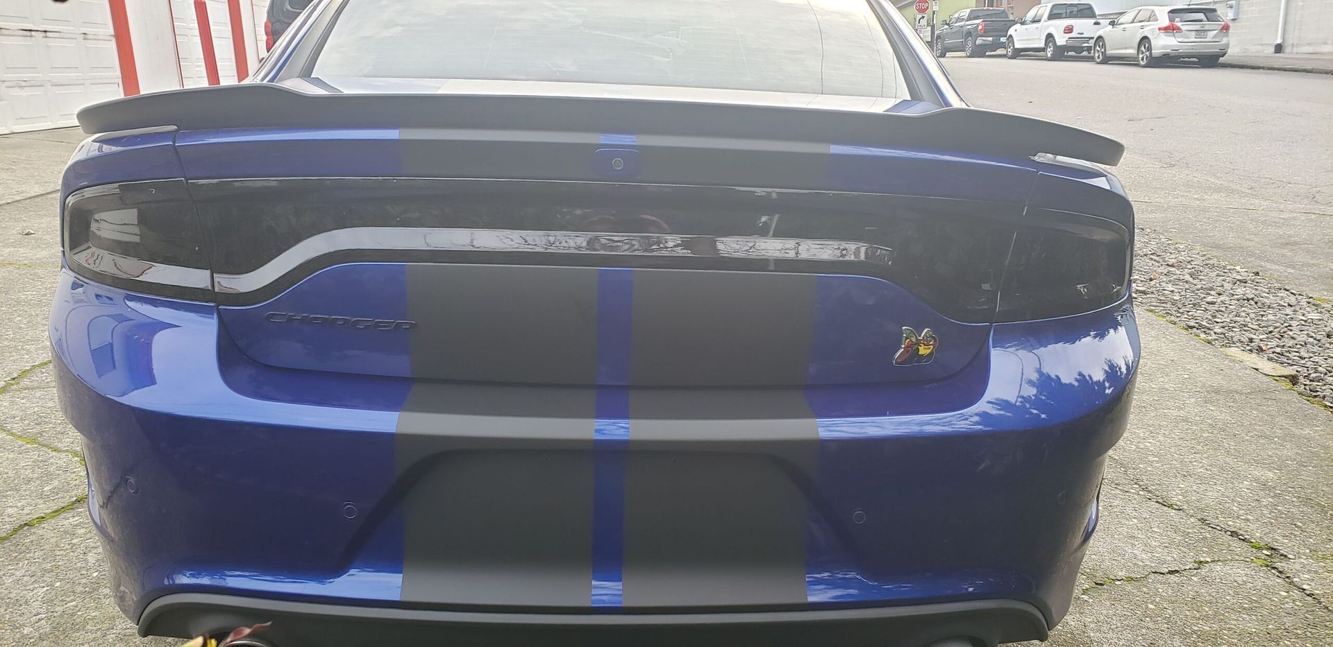 Blue sports car rear with black spoiler and twin racing stripes, parked outdoors.