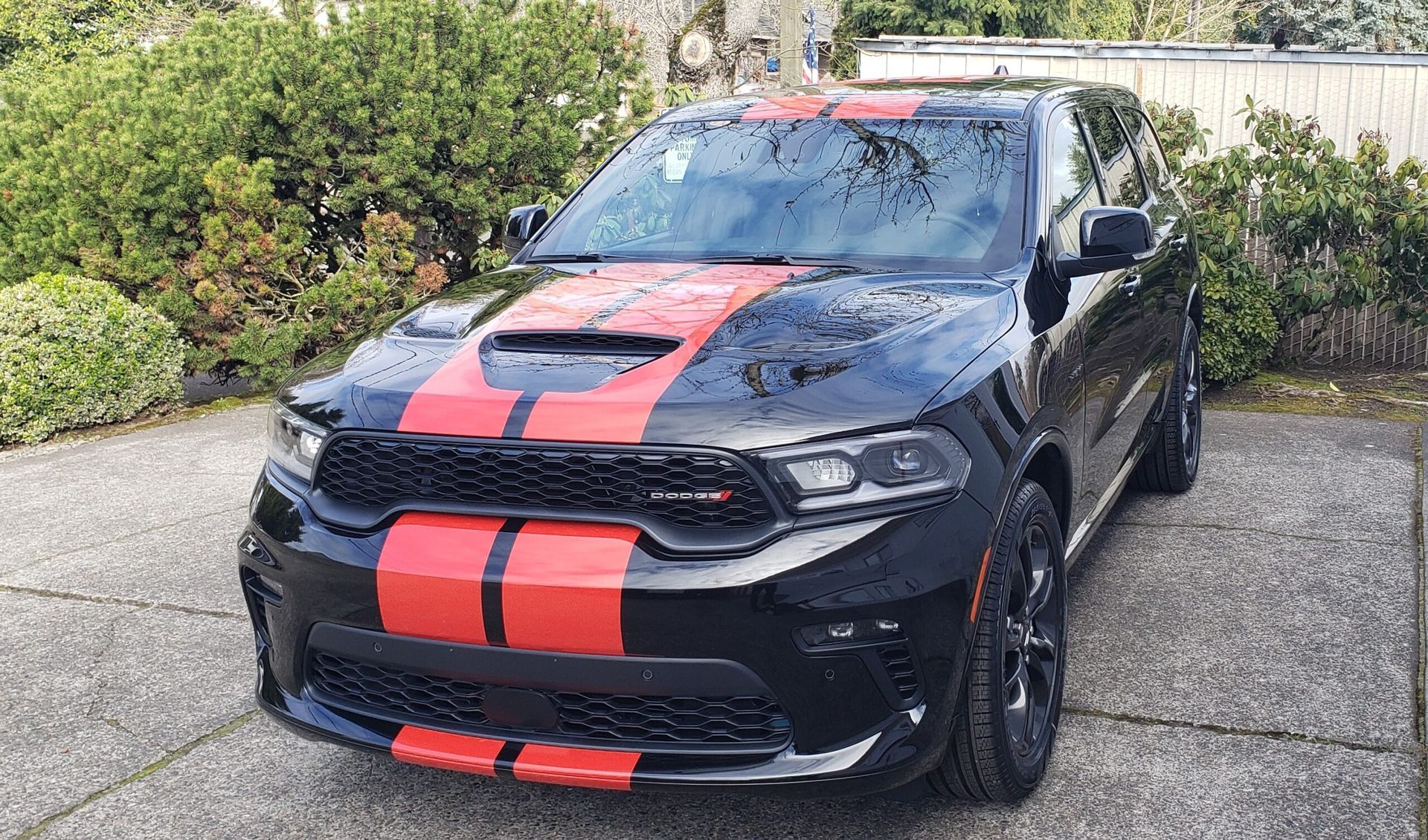 Black sports car with red racing stripes parked on a driveway, with bushes in the background