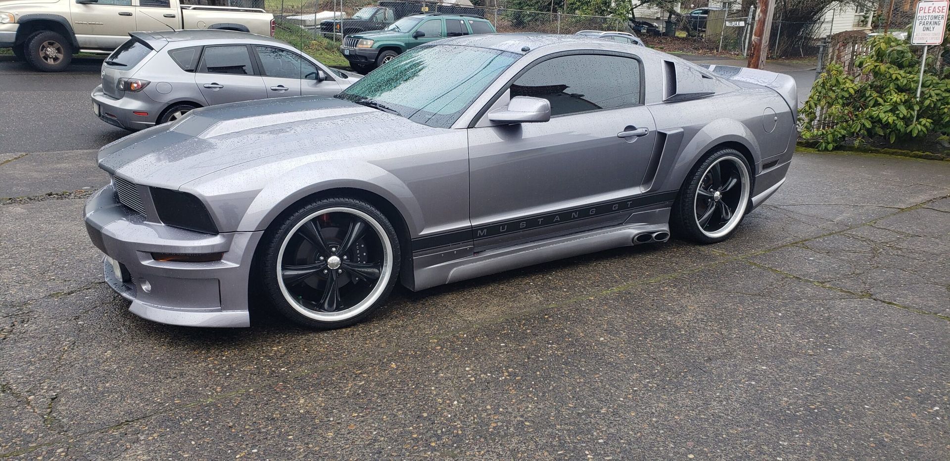 Silver sports coupe parked on a wet lot, side view with black wheels and body kit
