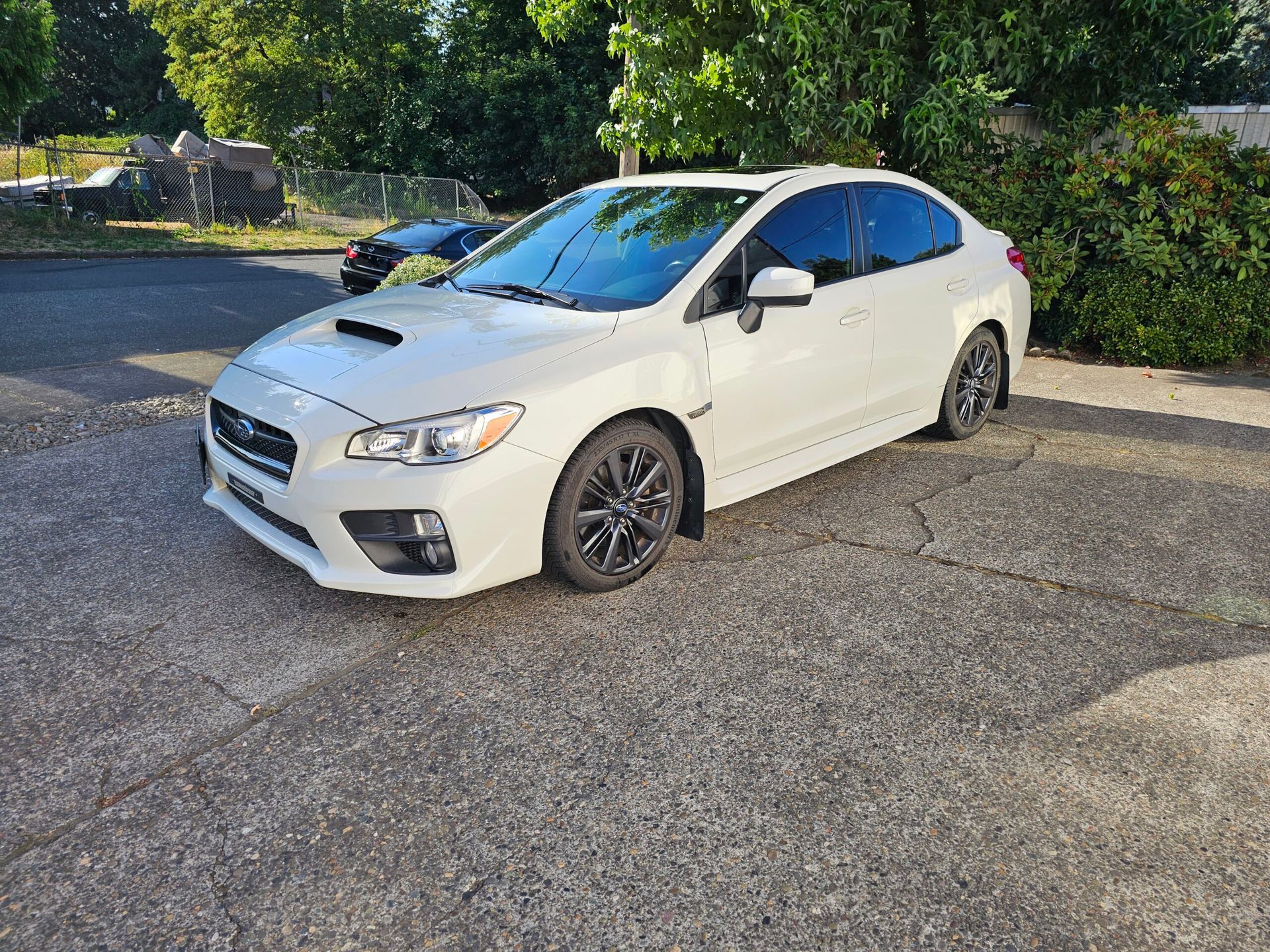 White sedan parked on a gravel lot beside trees, front-left view.