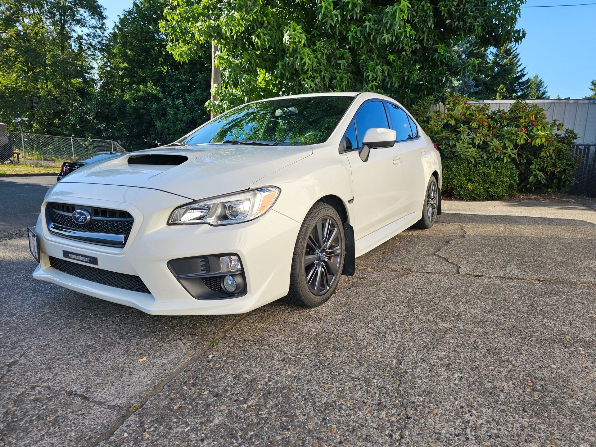 White Subaru sedan parked on gravel beside greenery, front-left view, sunny day