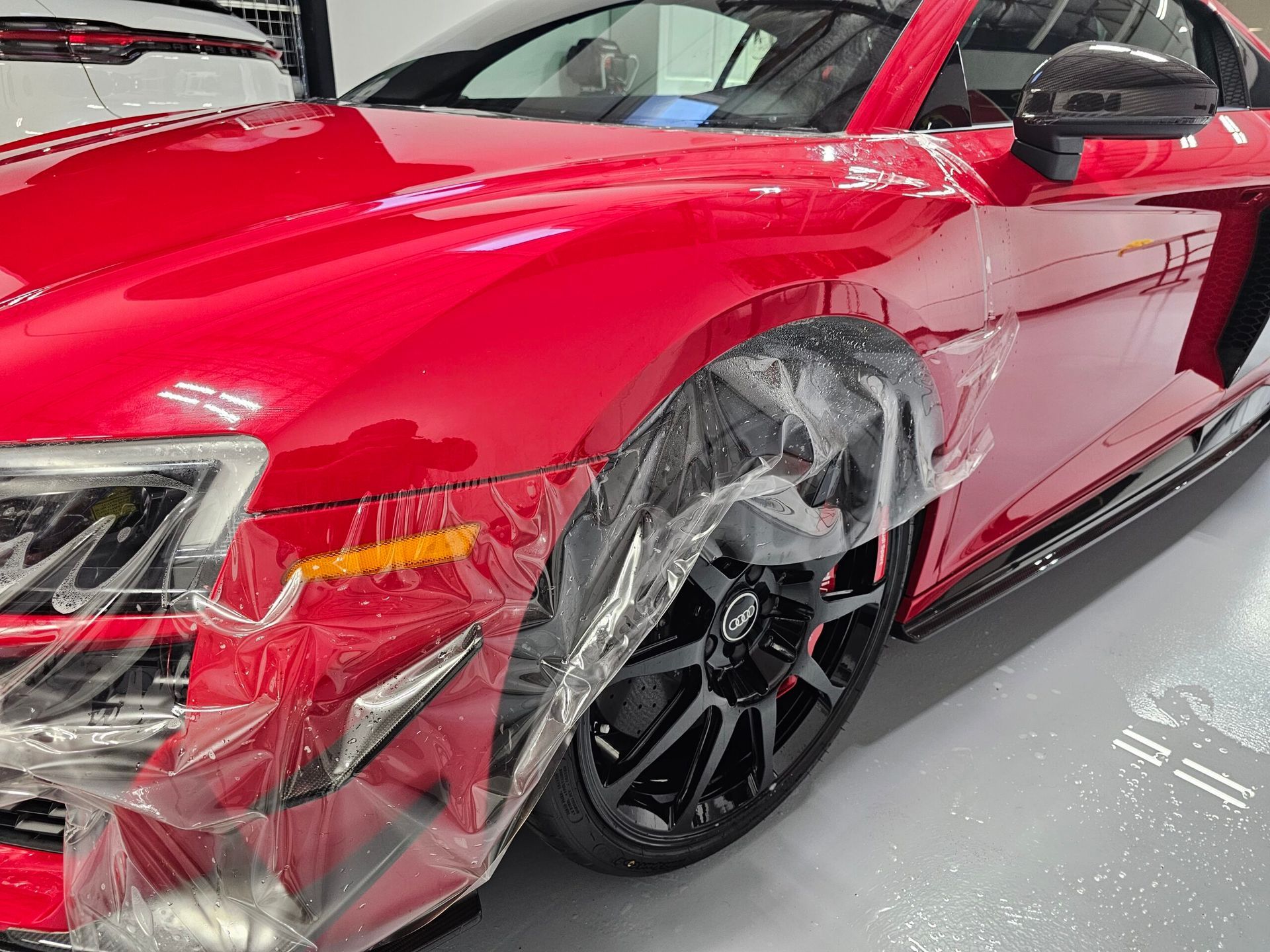 Red sports car with front-left damage, wrapped in clear plastic, in a garage.
