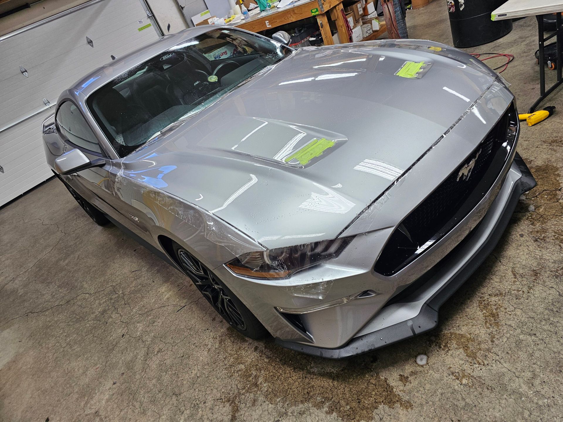 Silver sports car in a workshop, shown front-left with hood and front bumper removed.