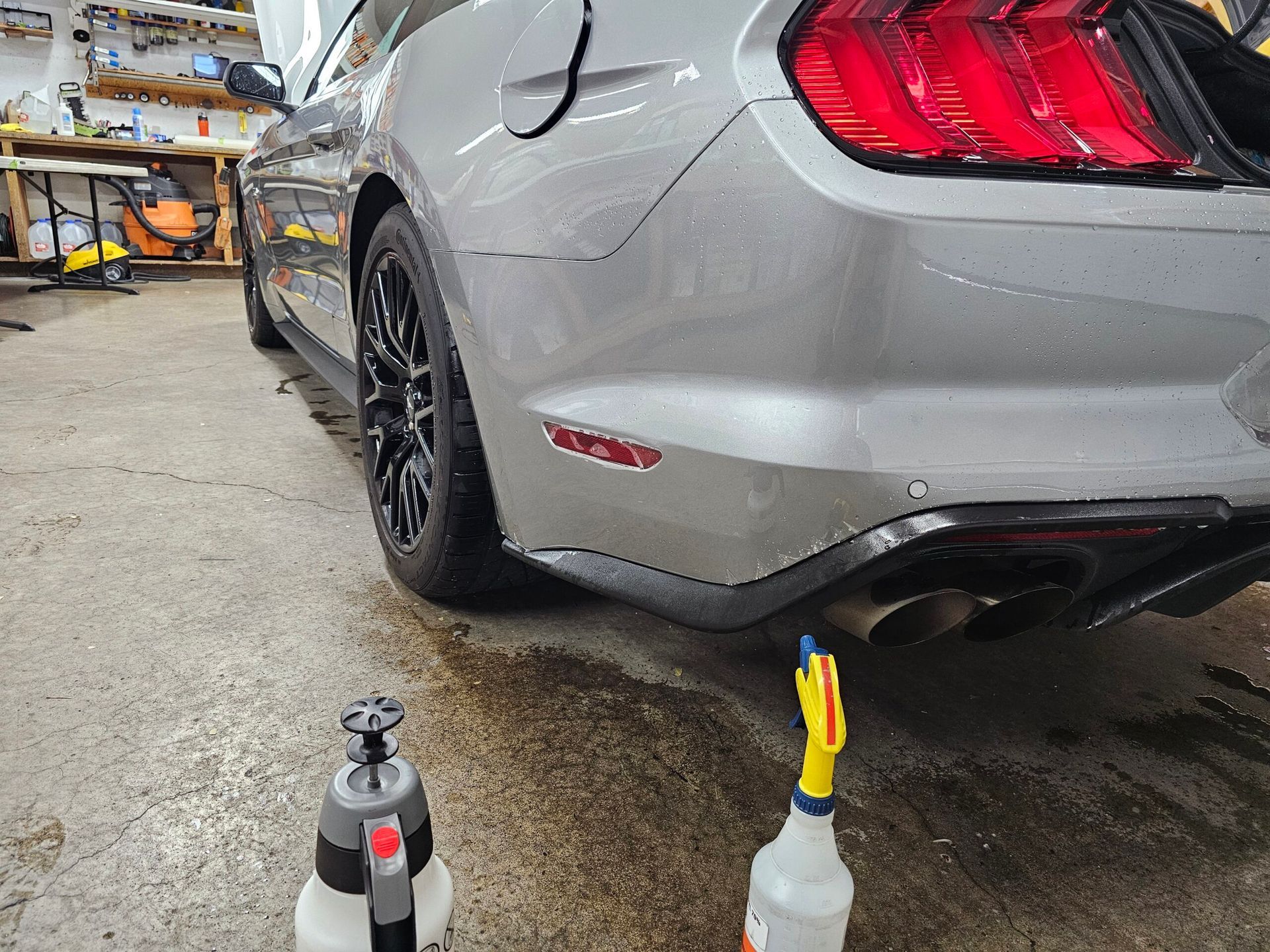 Silver sports car rear bumper being detailed in a garage, with spray bottles on the wet floor.