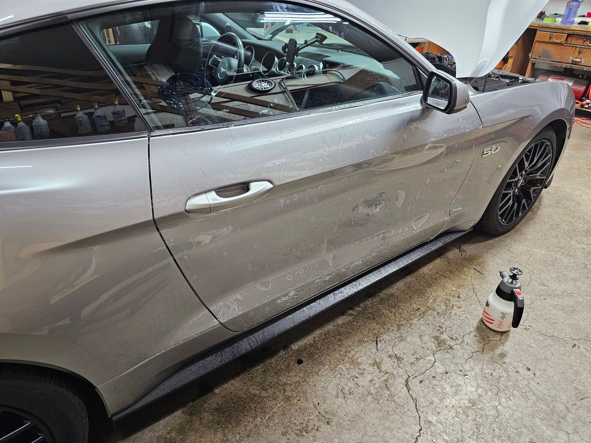 Silver car with side damage and broken rear window, parked indoors beside a spray bottle on the floor