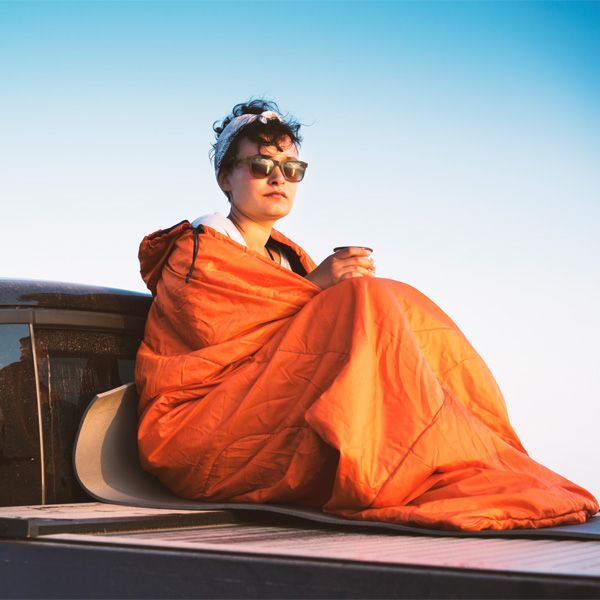 Person in sunglasses wrapped in an orange blanket sits on a rooftop under a clear blue sky.