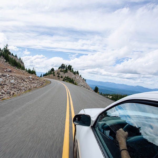 White car driving on a winding mountain road under a cloudy sky with distant hills.