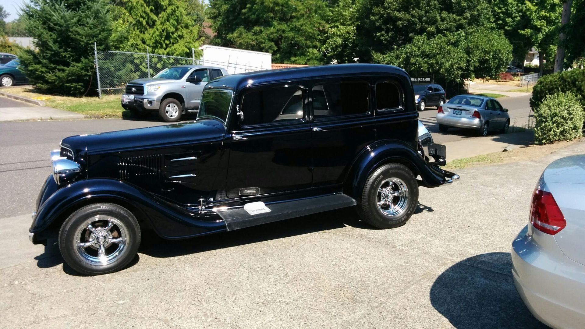 Black vintage coupe parked on a gravel lot with trees and cars in the background