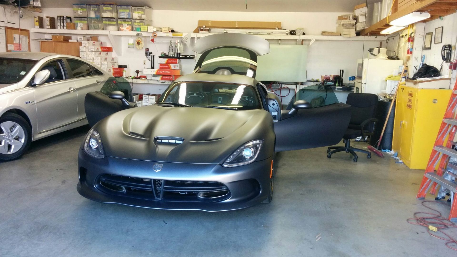 Dark sports car in a cluttered garage with its hood and trunk open and the driver door ajar