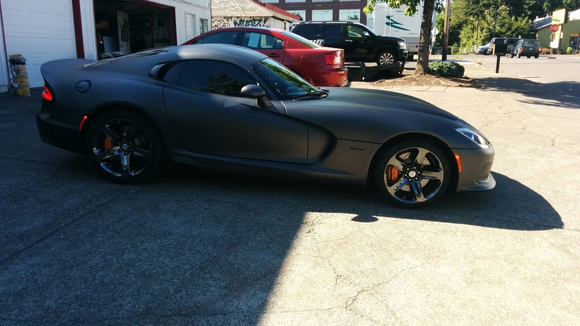 Matte black sports car parked outdoors in a sunny lot, side view with a red car behind it