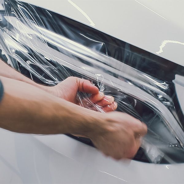 Hands peeling clear plastic wrap from a black car surface