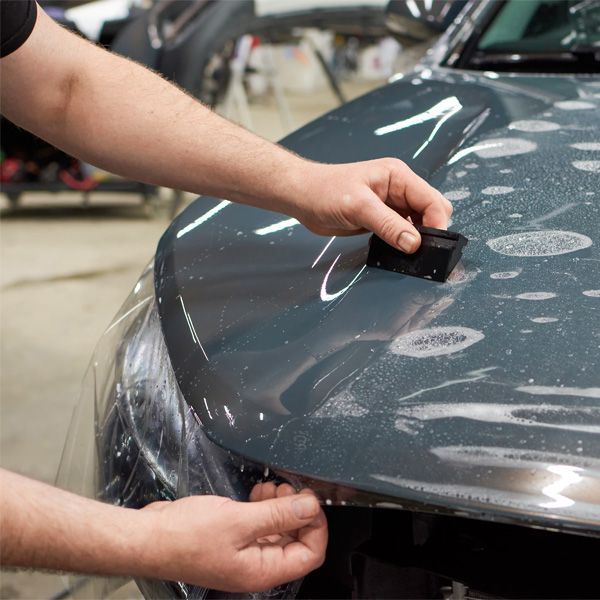 Hands applying vinyl wrap to a car hood with a small tool