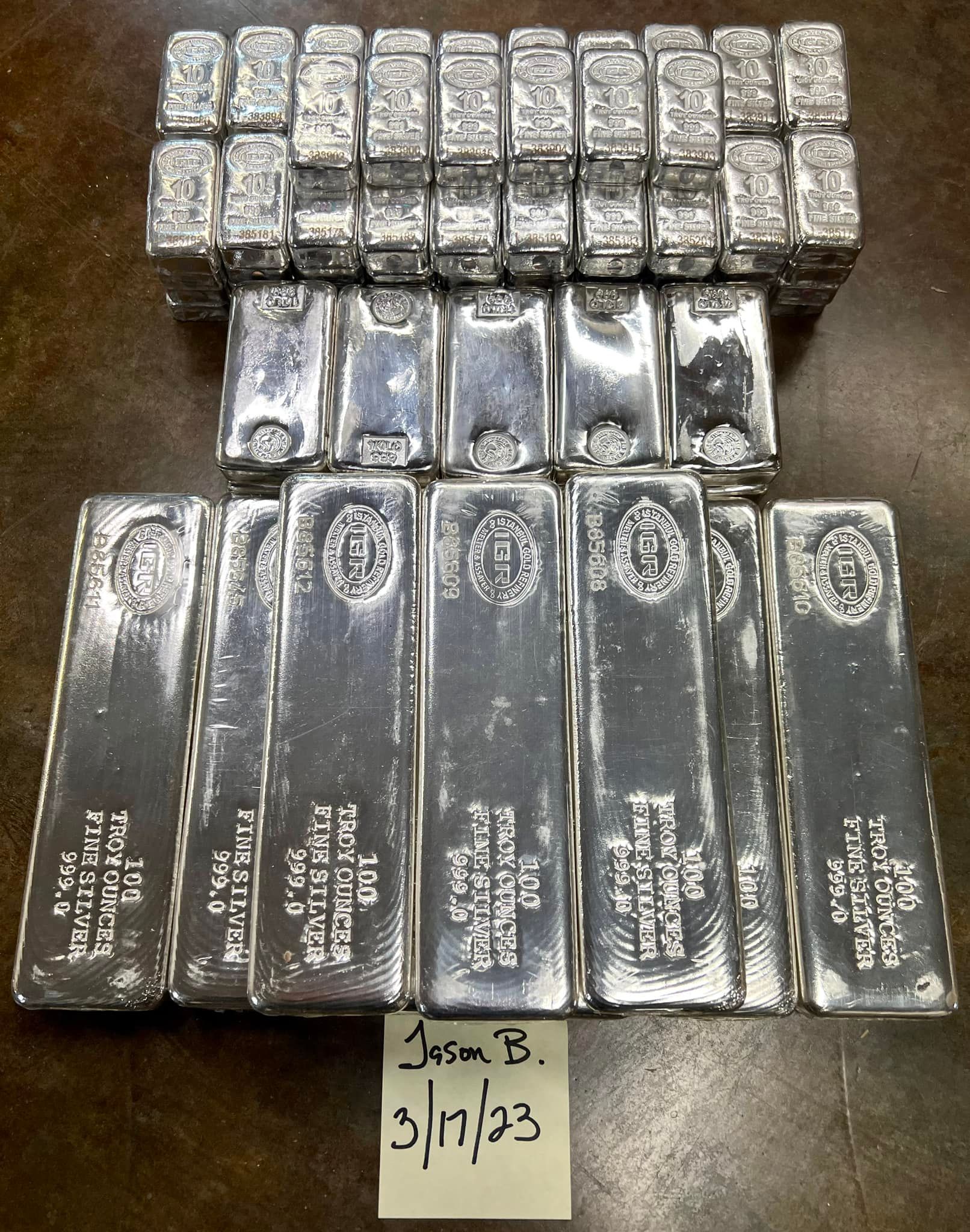 Rows of shiny silver metal bars stacked on a dark surface, with a small handwritten note below