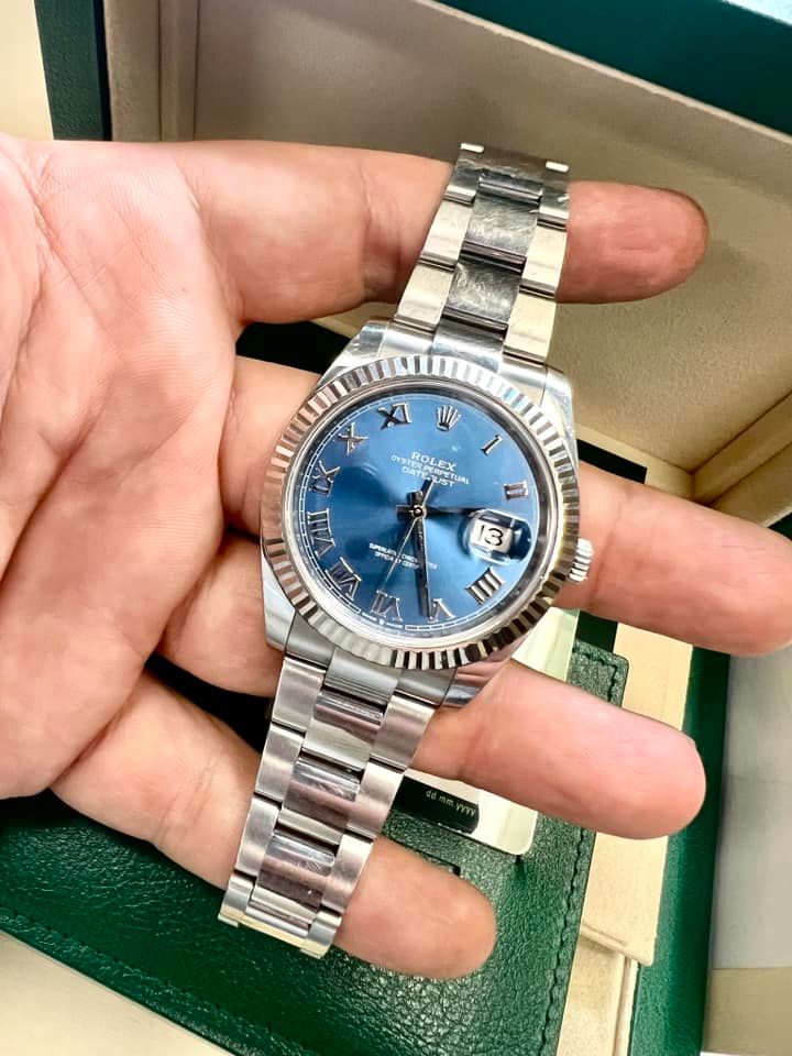 A person's hand holding a silver Rolex Datejust watch with a blue dial.