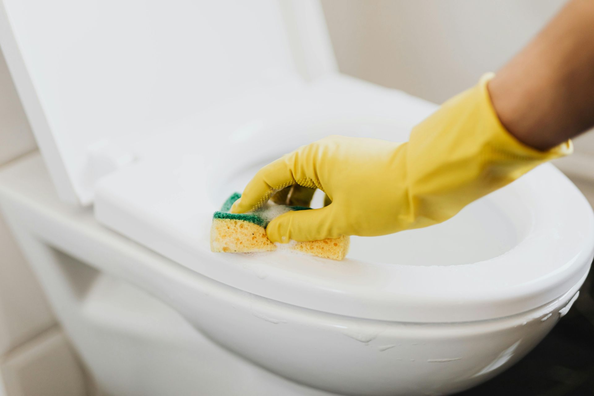Hand in yellow glove cleaning a white toilet seat with a sponge.