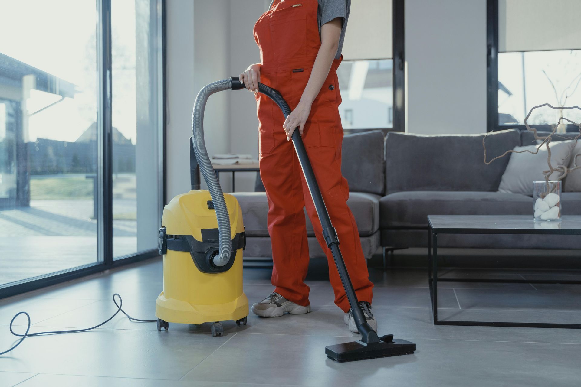 Person in orange jumpsuit vacuums a living room with large windows, yellow vacuum cleaner.