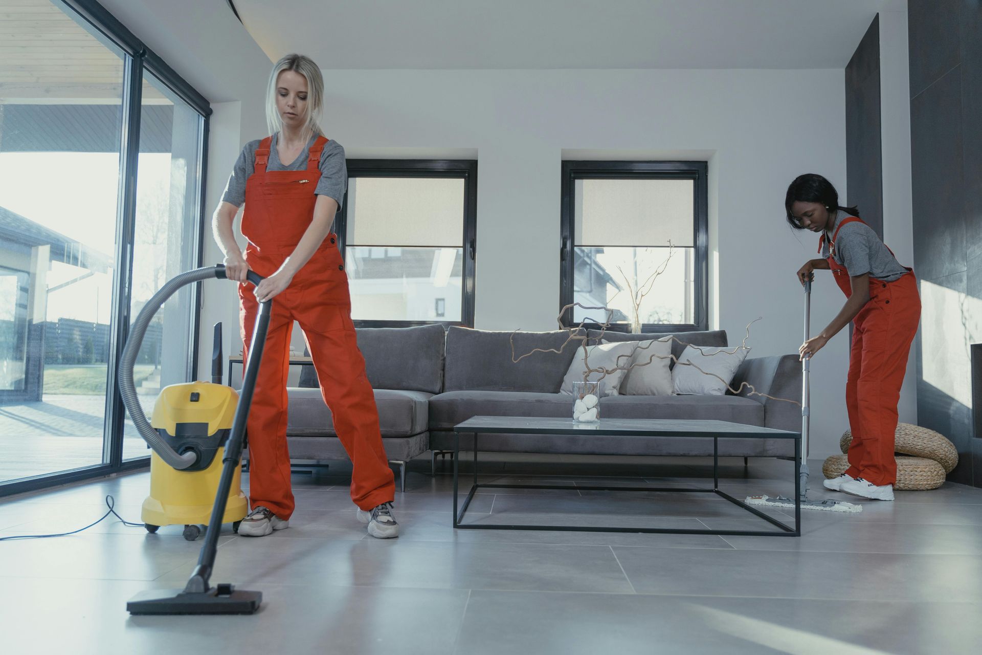 Two people cleaning a modern living room. One vacuums, the other mops. Both wear orange jumpsuits.