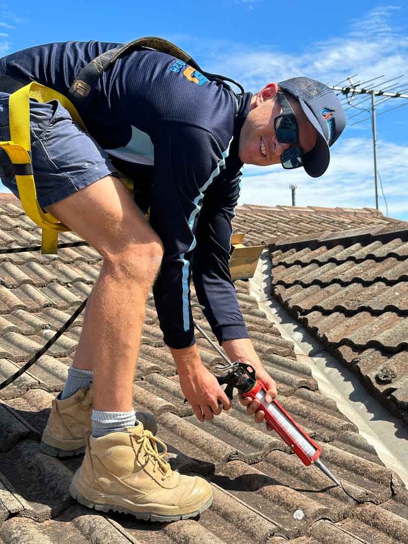 A Man is Kneeling on a Roof Holding a Bottle of Sealant — Wello's Plumbing & Gas In Thurgoona, NSW