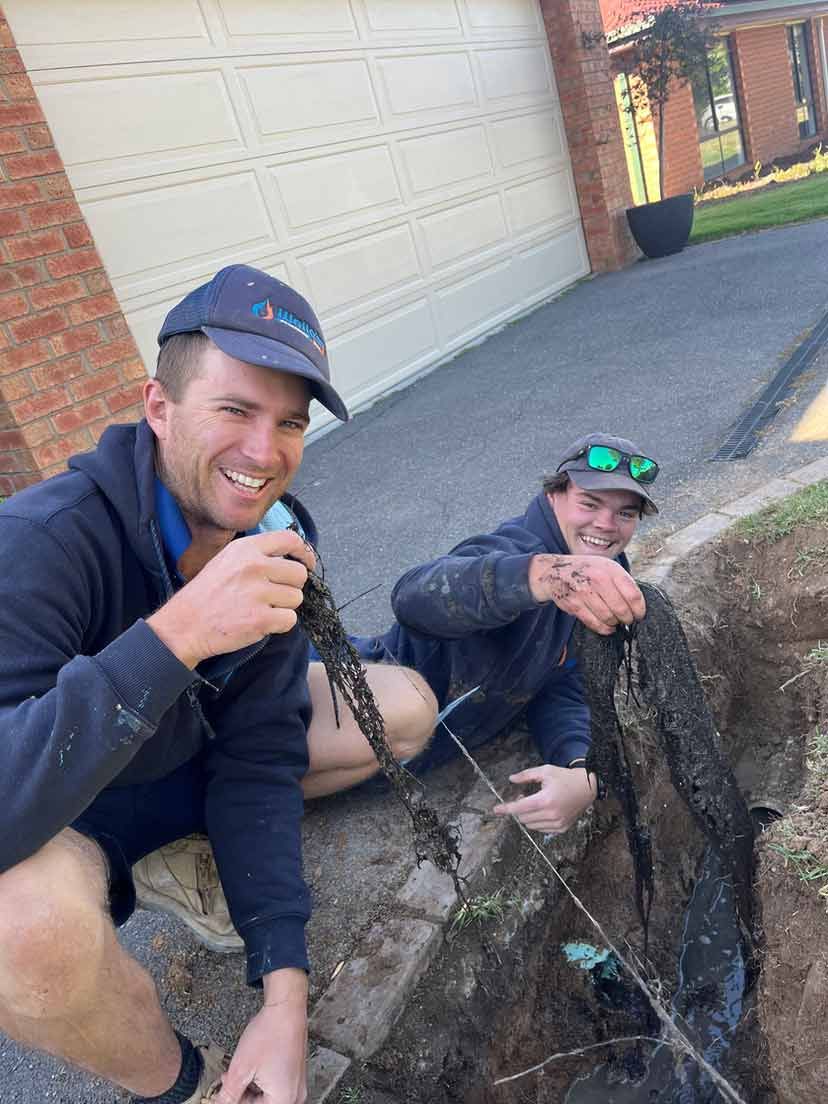 Two Men Are Kneeling Down in a Hole in the Ground — Wello's Plumbing & Gas In Thurgoona, NSW