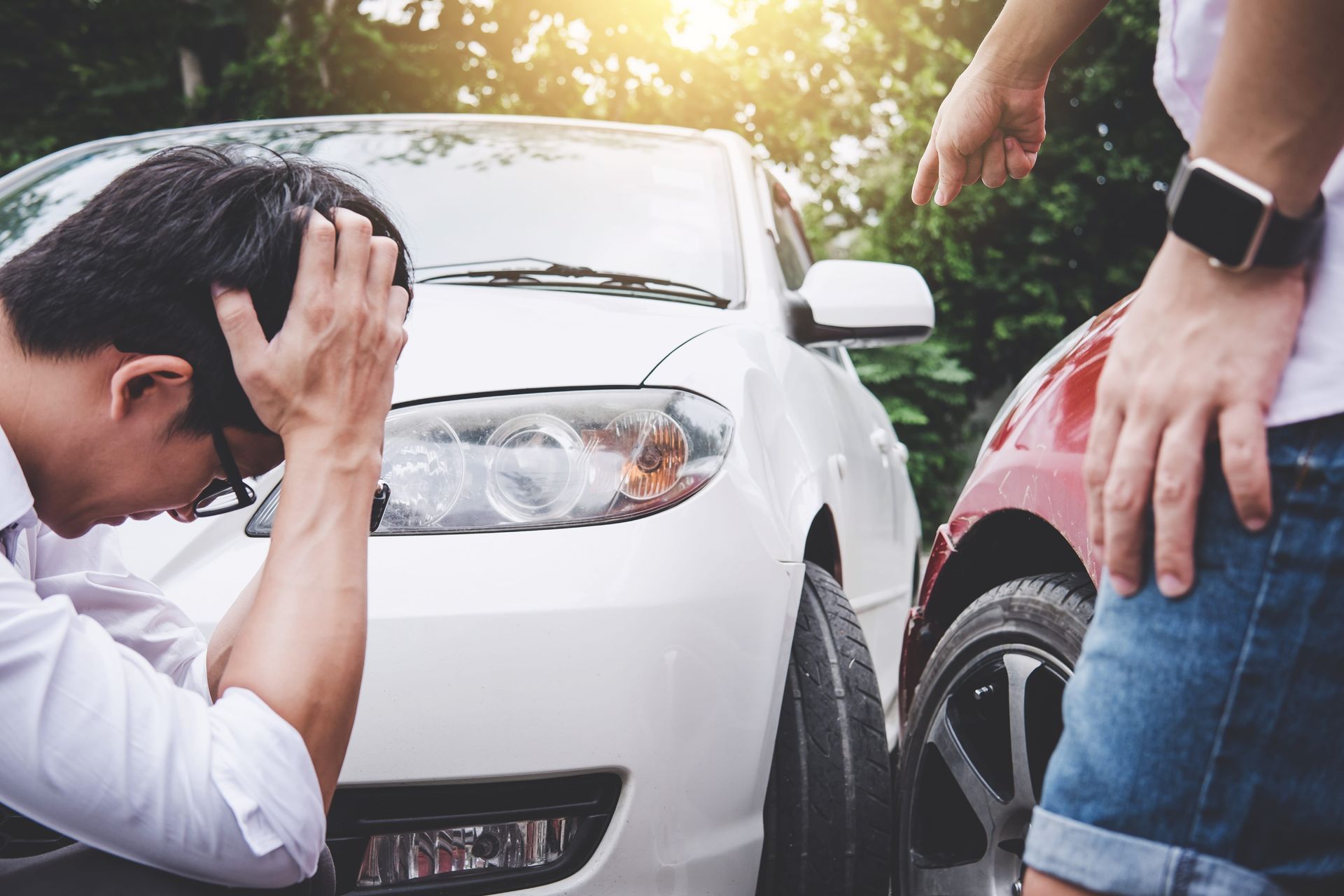 Man crouching beside damaged white car, holding his head, with another person nearby on the roadside