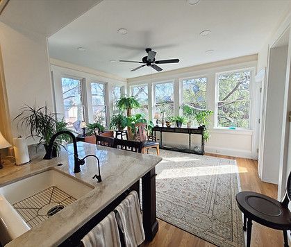 Bright sunroom with large windows, many potted plants, dining table, rug, and ceiling fan