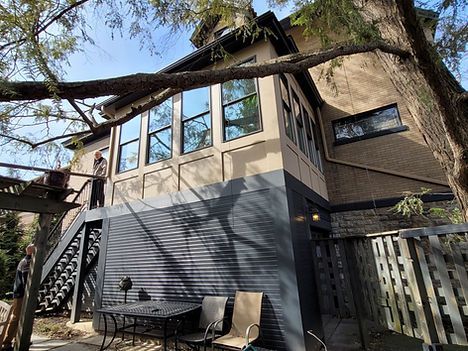 Modern house exterior with large windows, tree branches overhead, and patio furniture on a deck