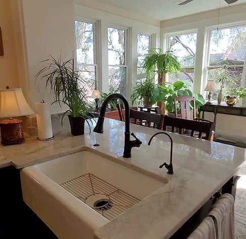 Bright kitchen with a marble island sink, black faucet, and large windows with potted plants.
